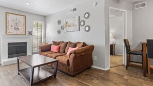 A living room with a brown sofa, a coffee table, and a fireplace. There are wall decorations and a door leading to another room. - WORK REMOTELY IN URBAN DFW! (Dallas)