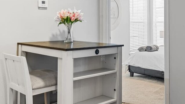 Una piccola scrivania bianca in legno, con un vaso di fiori, una sedia e un letto con asciugamani arrotolati sullo sfondo.