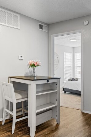 Un petit bureau en bois blanc, sur lequel se trouve un vase de fleurs, une chaise, et en arrière-plan, un lit avec des serviettes pliées.