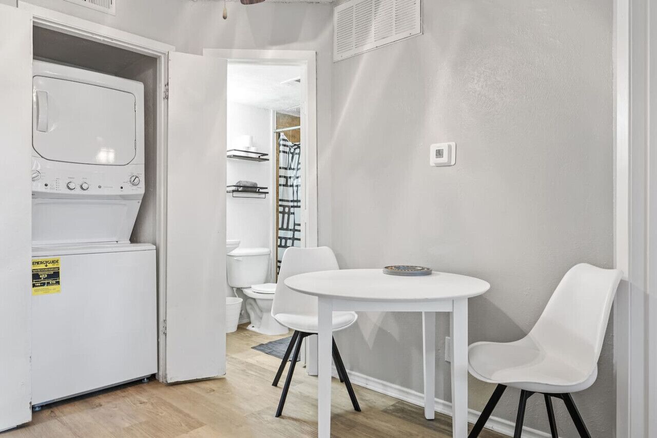 Cocina compacta con lavadora y secadora integradas, una pequeña mesa de comedor con sillas, y un baño a la vista al fondo.
