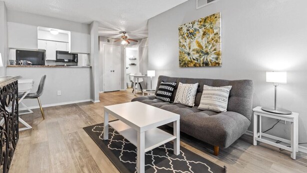 A modern living room with a grey sofa, white coffee table, and a black and white rug. A kitchen area is visible in the background with white cabinets and a microwave. - Affordable & Convenient! Free WiFi! (Dallas)