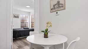 A small, white-walled room with a round table, two chairs, a single orchid in a glass vase, and a bed with a dark cover in the background. - Cute & Contemporary 1 Bed Apt! Easy Pool Access! (Dallas)