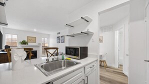A modern kitchen with a stainless steel sink, a microwave, and a dishwasher. There is a dining area with a white table and chairs. - Cute & Contemporary 1 Bed Apt! Easy Pool Access! (Dallas)
