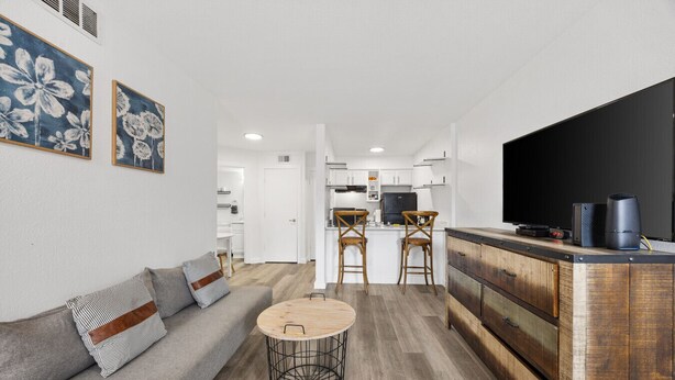 A modern living room with a grey sofa, a round wooden coffee table, and a kitchen area with white cabinets and a black refrigerator. - Cute & Contemporary 1 Bed Apt! Easy Pool Access! (Dallas)