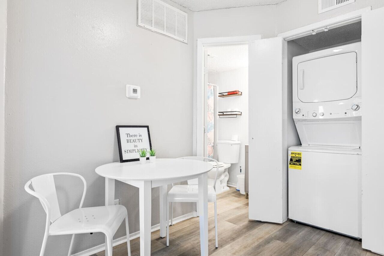 Una habitación pequeña y limpia, con una mesa y sillas blancas, una lavadora y una puerta que da a otra habitación.