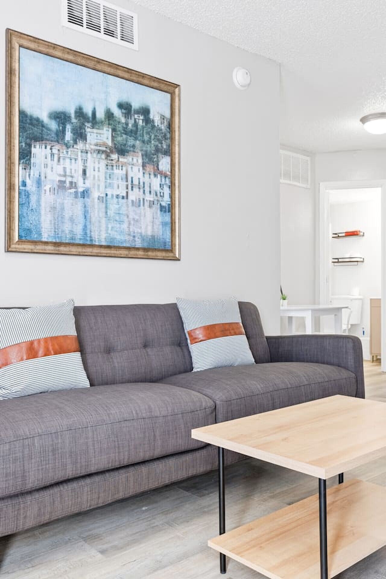 Een moderne woonkamer met een grijze bank, gestreepte kussens, een houten salontafel en een groot schilderij van een kustplaatsje.