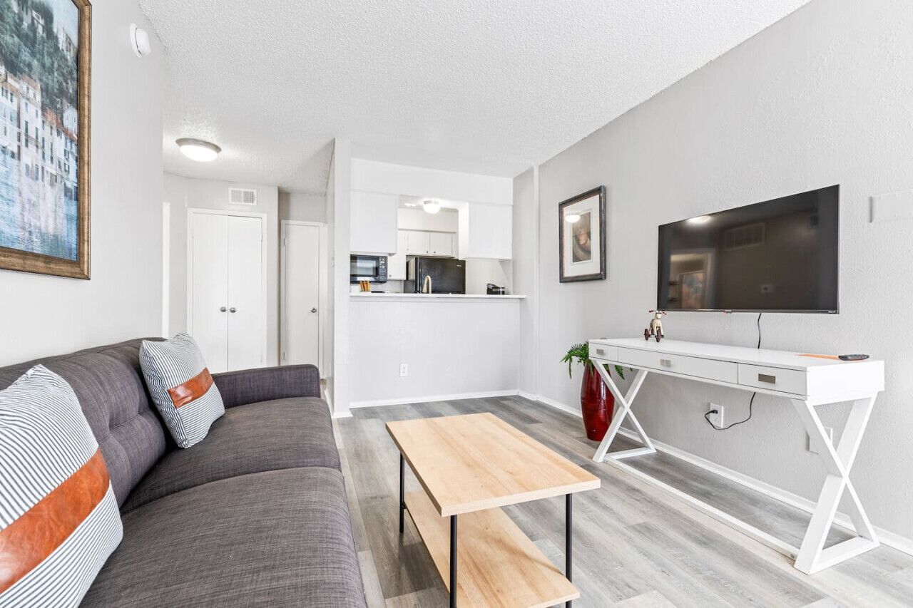 A modern living room with a gray sofa, a wooden coffee table, a white desk, and a flat-screen TV mounted on the wall.