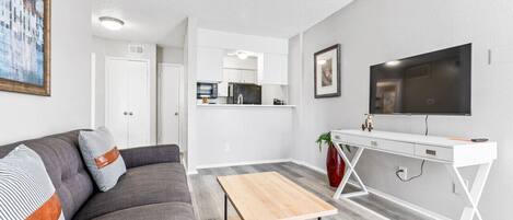 A modern living room with a gray sofa, a wooden coffee table, a white desk, and a flat-screen TV mounted on the wall.