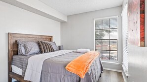 Een slaapkamer met een houten bedframe, grijze beddengoed en een raam met zonwering.