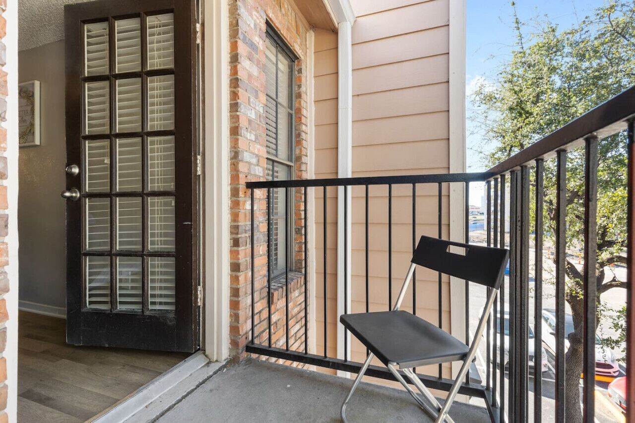 A black door with a window, a brick wall, a beige siding building, a black metal railing, and a black folding chair on a balcony.