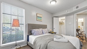 A bedroom with a bed, a bedside table, a lamp, a window with blinds, and a door leading to a bathroom. - Central DFW 1 Bed Apt! (Dallas)