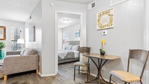 A modern living room with a sofa, a dining table, and chairs. There is a bed in the background, and a wall with a decorative frame. - Central DFW 1 Bed Apt! (Dallas)