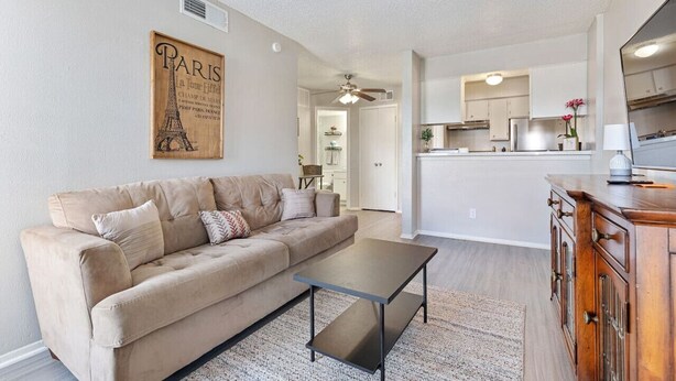 Un salon avec un canapé beige, une table basse et un meuble en bois. Au fond, on aperçoit une cuisine avec des meubles blancs et un ventilateur de plafond.