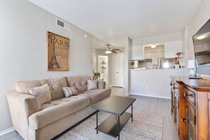 Una sala de estar con un sofá color beige, una mesa de centro y un aparador de madera. Se observa una cocina al fondo con armarios blancos y un ventilador de techo.