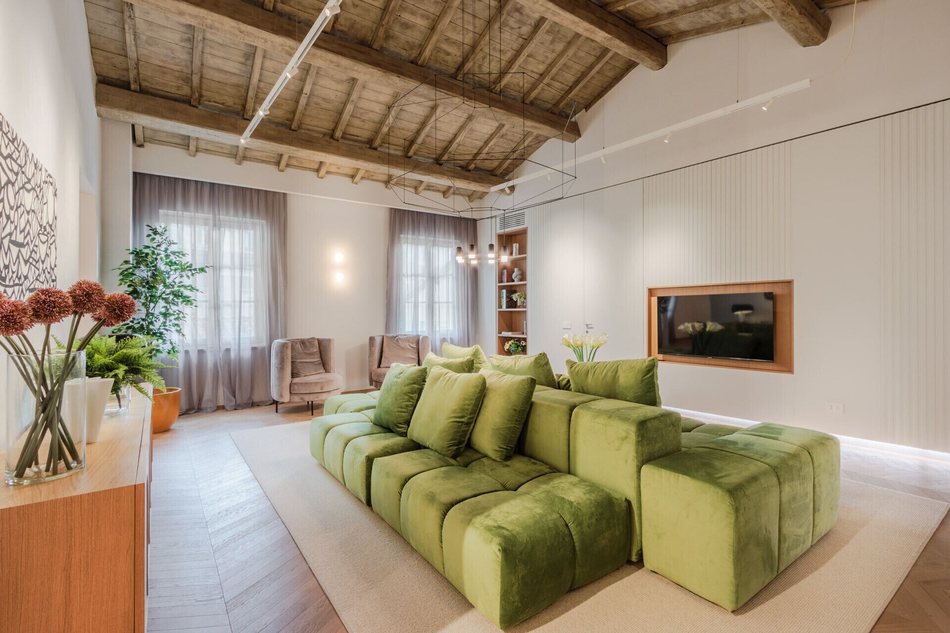 Un salon moderne avec un canapé d’angle vert, des poutres en bois au plafond et un téléviseur à écran plat fixé au mur.