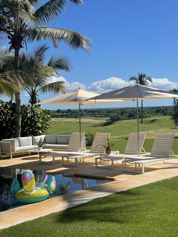 A poolside area with lounge chairs, umbrellas, and a view of a golf course.