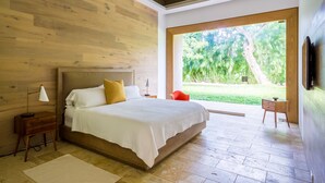 A modern bedroom with a wooden headboard, a large bed with white bedding, a nightstand, a lamp, and a view of a tropical garden through a large window.