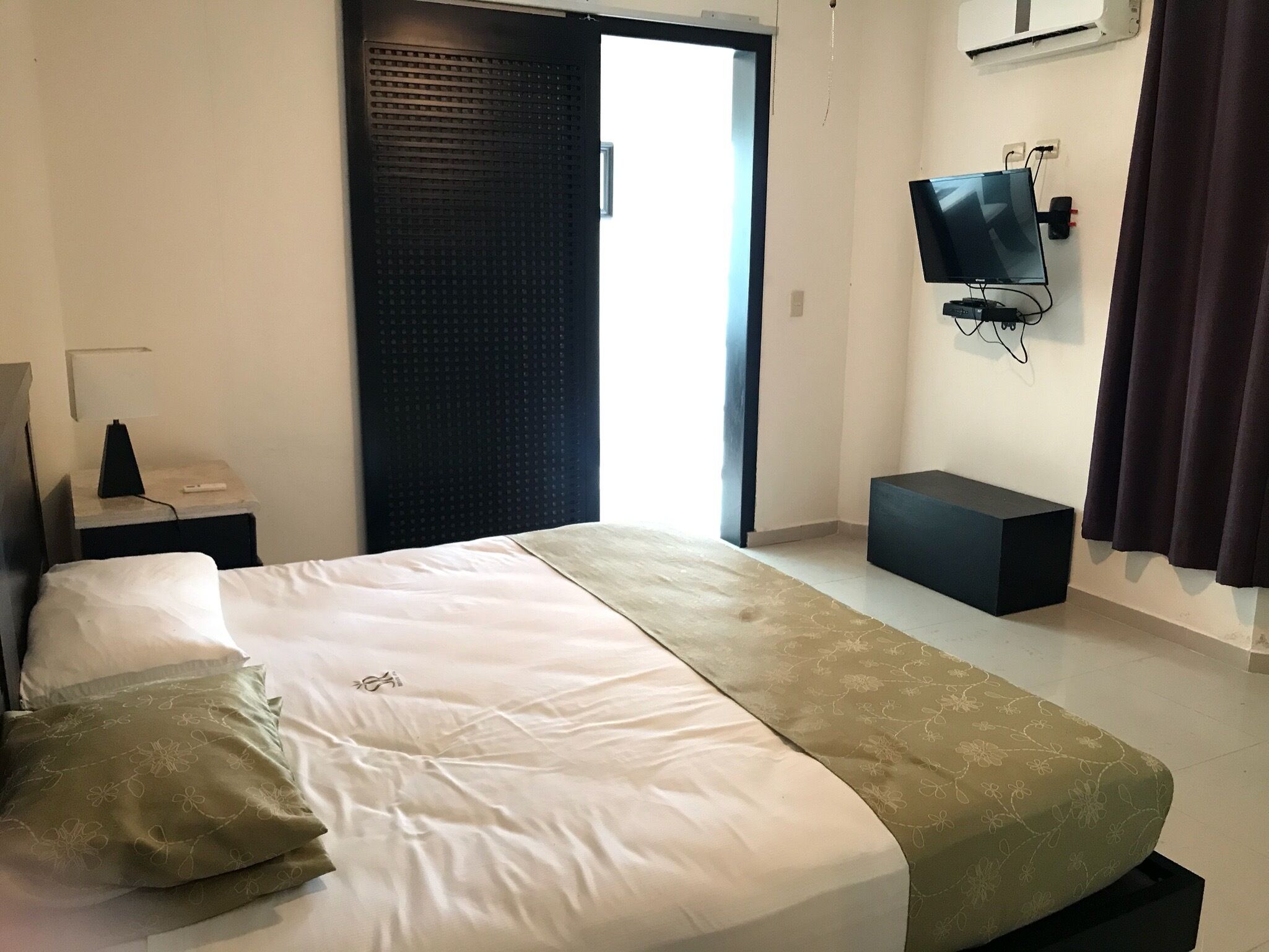 Een hotelkamer met een bed, een aan de muur bevestigde televisie, een bureau met een lamp en een raam met gordijnen.