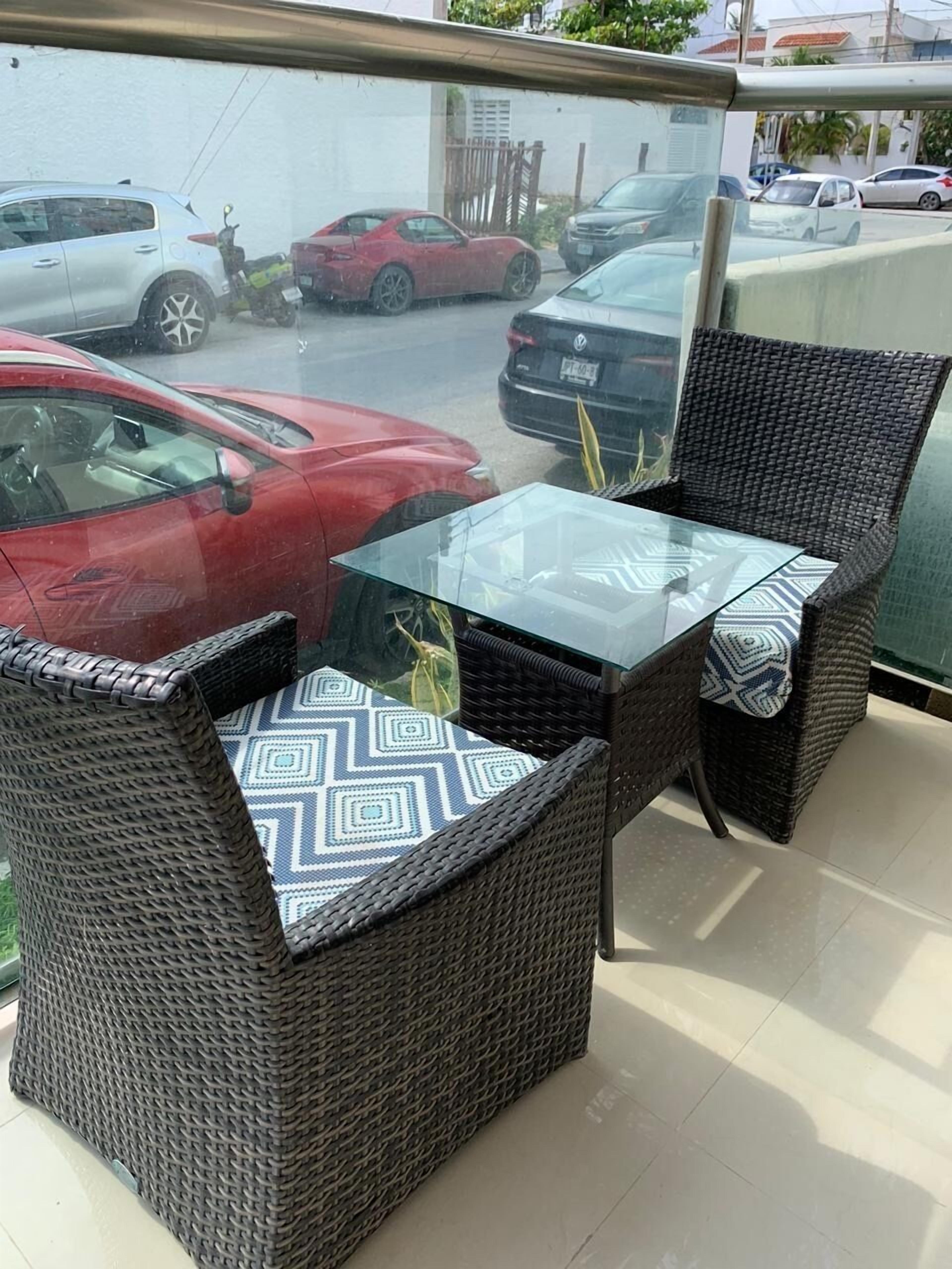 Un balcón con mobiliario de mimbre y una mesa de vidrio, con vista a una calle con coches estacionados.