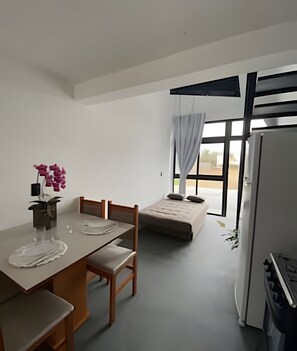 A small, modern room with a dining table, chairs, a sofa, a refrigerator, and a window with curtains. - Lofts Meia Morada (São Francisco do Sul)
