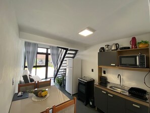 A compact kitchen with a microwave, stove, and sink. There is a dining table with fruit and a remote control. A staircase leads to an outdoor area with a view of a building. - Lofts Meia Morada (São Francisco do Sul)