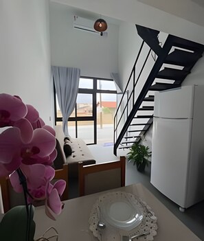 A modern kitchen with a built-in refrigerator, a dining area with a table and chairs, a staircase, and a large window with curtains. - Lofts Meia Morada (São Francisco do Sul)