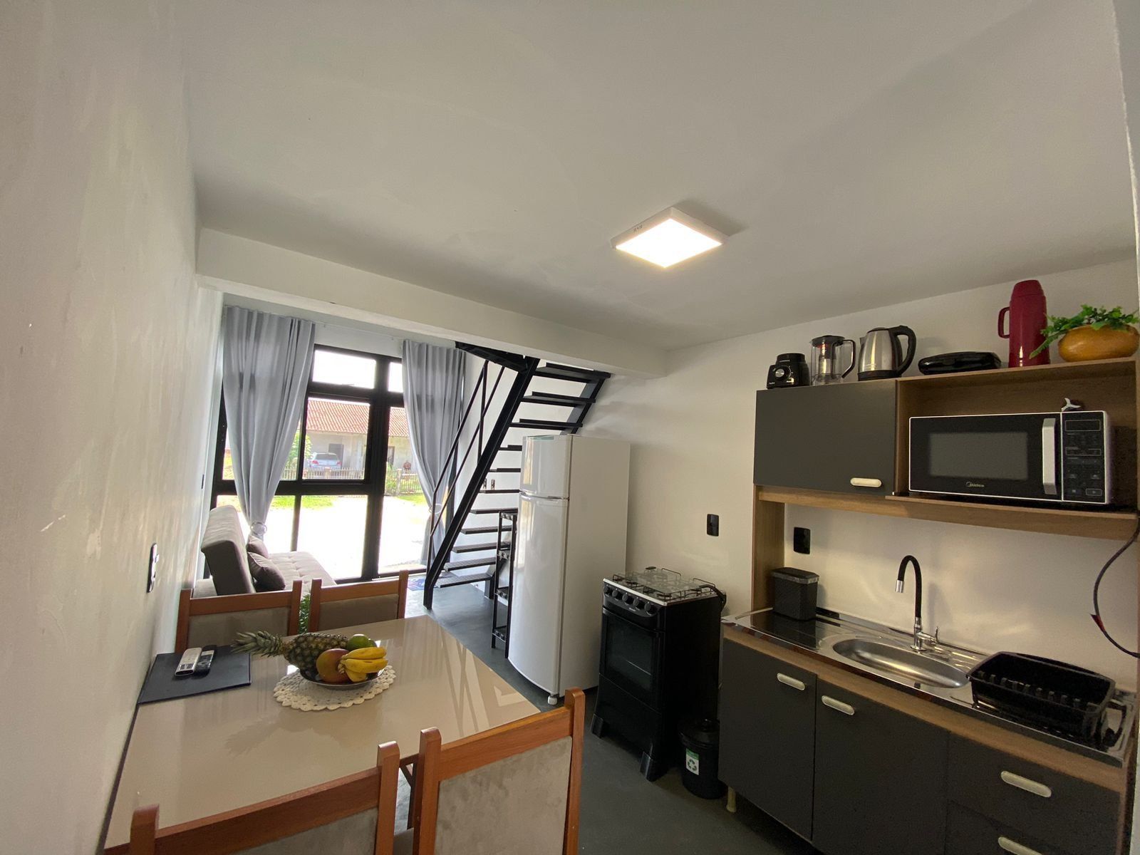 A compact kitchen with a microwave, stove, and sink. There's a dining table with chairs and a small fruit bowl. A staircase leads to an outdoor area.