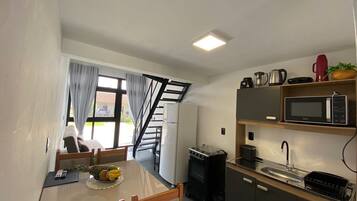 A compact kitchen with a microwave, stove, and sink. There's a dining table with chairs and a small fruit bowl. A staircase leads to an outdoor area.