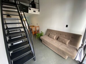 A staircase with a metal railing, a beige sofa, and a small table with chairs in a room with white walls. - Lofts Meia Morada (São Francisco do Sul)