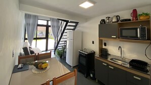 A compact kitchen with a microwave, stove, and sink. There is a dining table with fruit and a remote control. A staircase leads to an outdoor area with a view of a building. - Lofts Meia Morada (São Francisco do Sul)