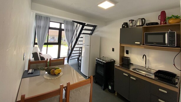 A compact kitchen with a microwave, stove, and sink. There's a dining table with chairs and a small fruit bowl. A staircase leads to an outdoor area. - Lofts Meia Morada (São Francisco do Sul)
