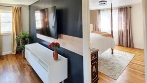 A modern bedroom with a large bed, a TV mounted on the wall, a white console table, and wooden flooring. - Chic, Spacious Pet Friendly Apt Near Attractions/Food/EWR/NYC (West Orange)