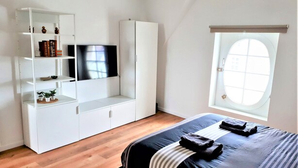 Une chambre à coucher avec un lit, un téléviseur posé sur un meuble blanc, une fenêtre et un plancher en bois.