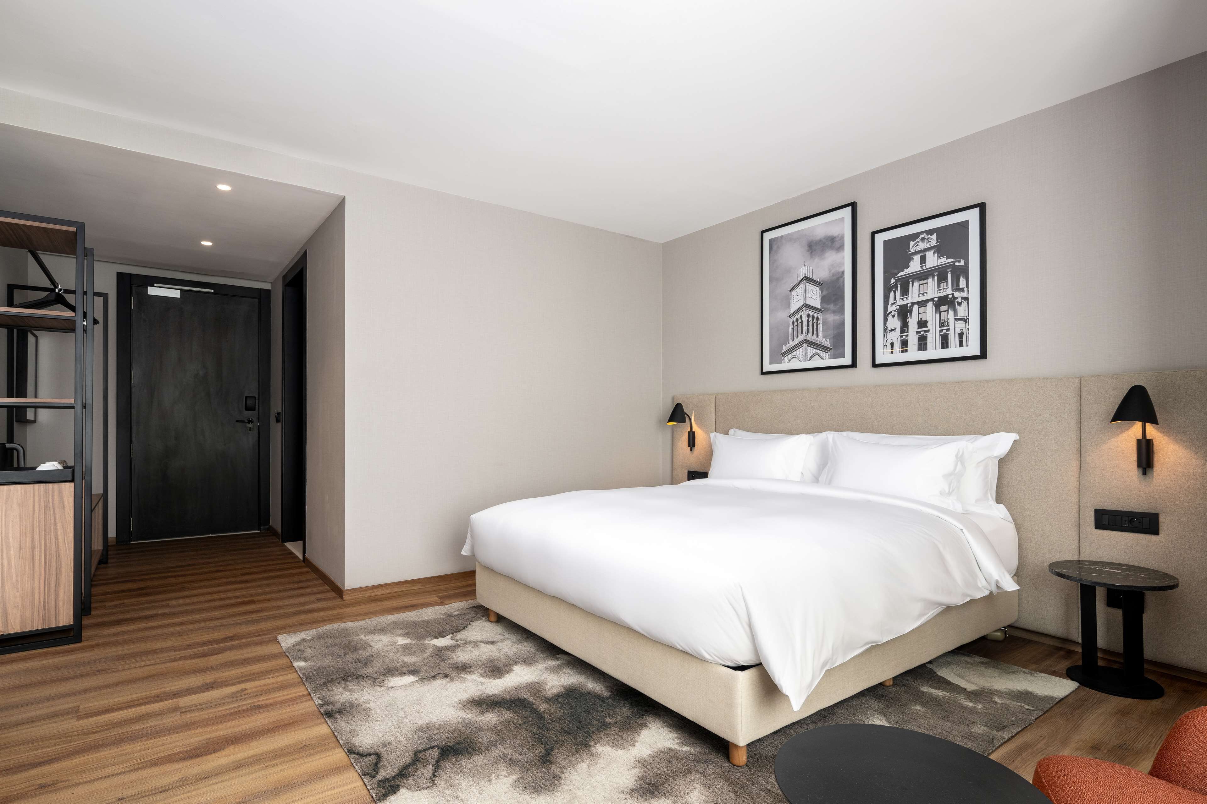A modern hotel room with a large bed, a desk, and a small table. There are two framed pictures on the wall.