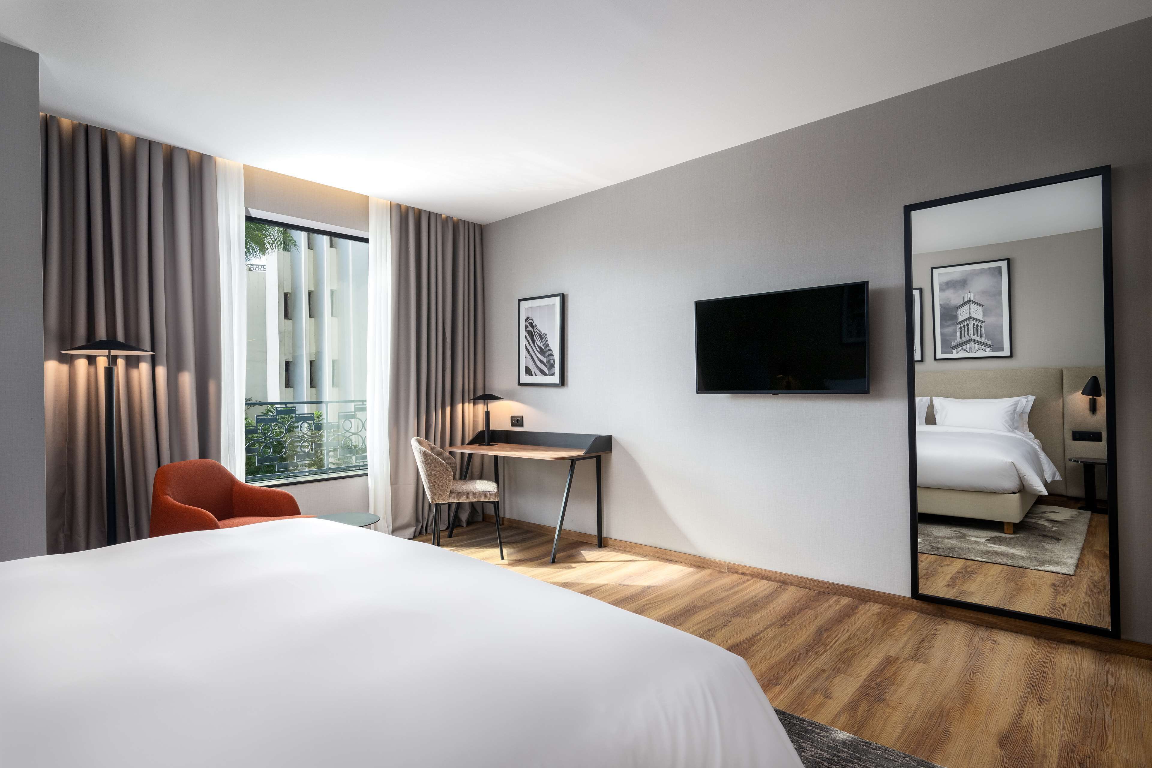 A modern hotel room with a large bed, a desk, a chair, a TV, and a large mirror.