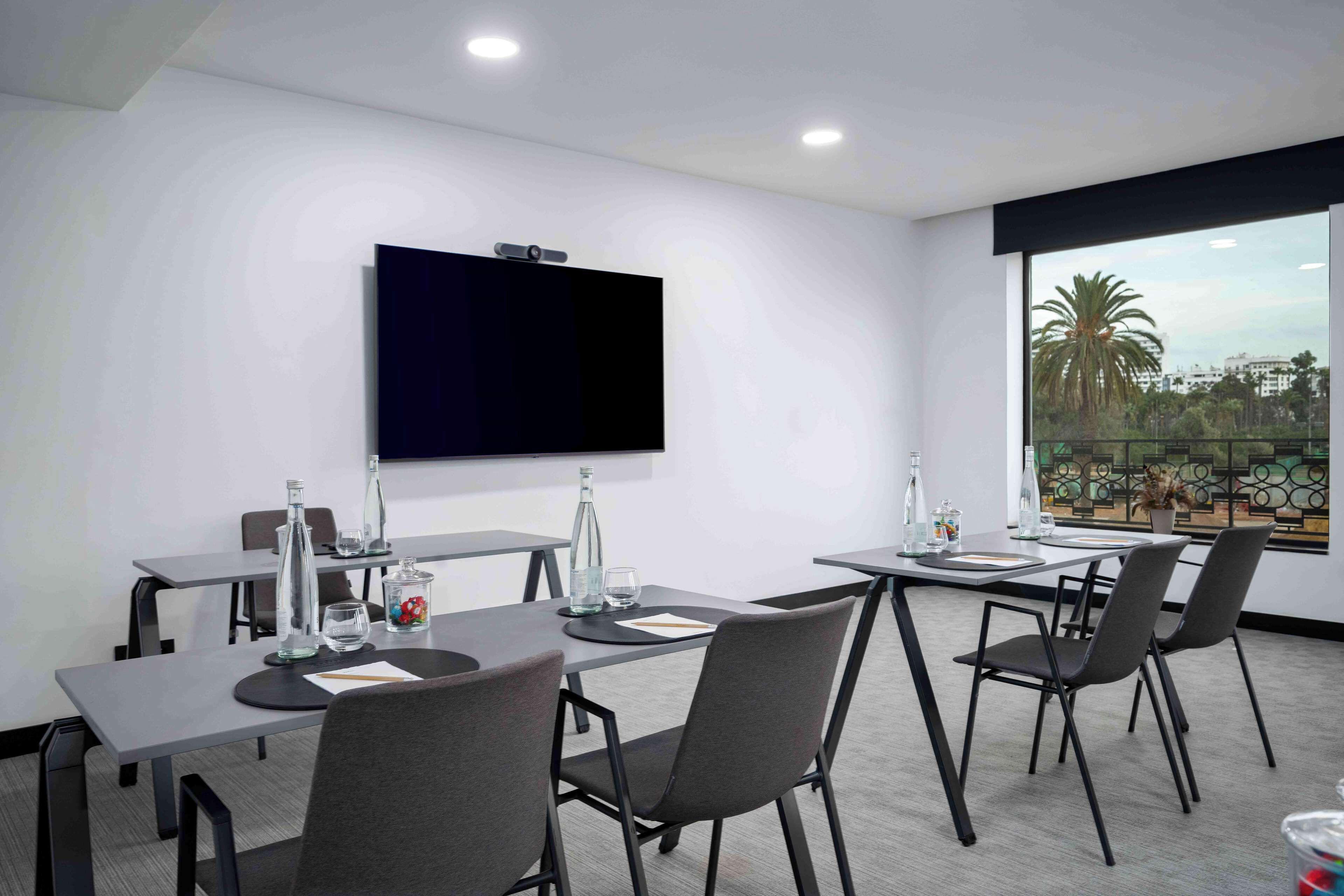 A modern conference room with a large flat-screen TV mounted on the wall, two tables with chairs, and a large window offering a view of palm trees and buildings.
