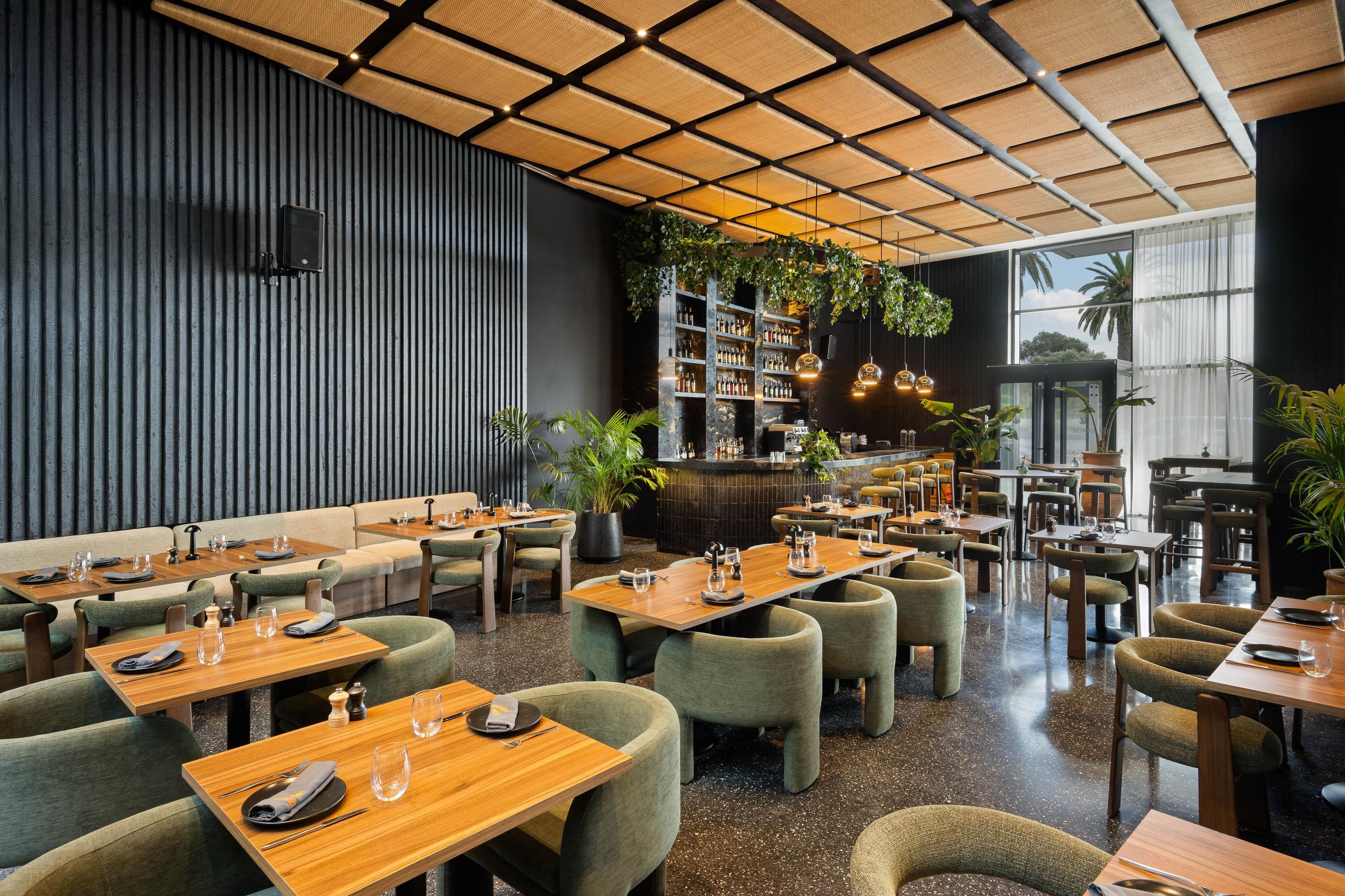 Un restaurante con varias mesas y sillas, una zona de bar con estantes llenos de botellas y un techo con diseño geométrico.