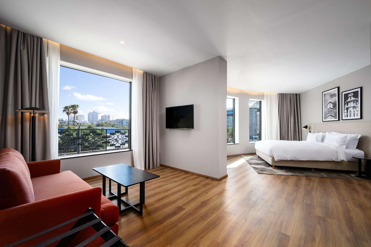 Une chambre d’hôtel moderne avec une grande fenêtre, un lit, un canapé rouge, une petite table et un téléviseur fixé au mur.