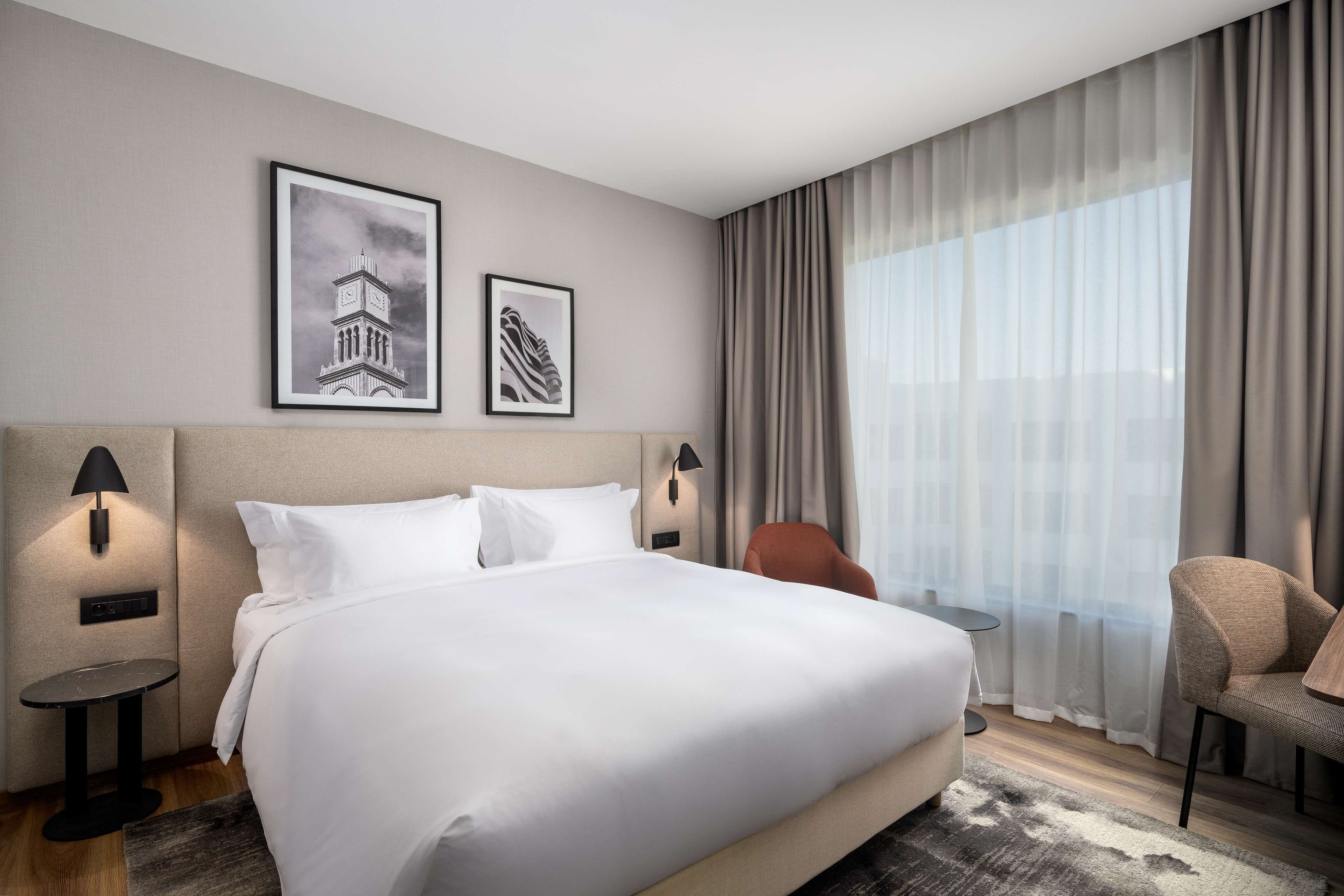 A hotel room with a large bed, two wall-mounted lamps, a small table, a chair, and a large window with curtains.
