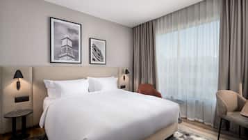 A hotel room with a large bed, two wall-mounted lamps, a small table, a chair, and a large window with curtains.
