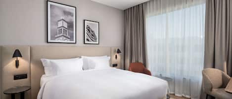 Ein Hotelzimmer mit einem großen Bett, zwei Wandlampen, einem kleinen Tisch, einem Sessel und einem großen Fenster mit Vorhängen.