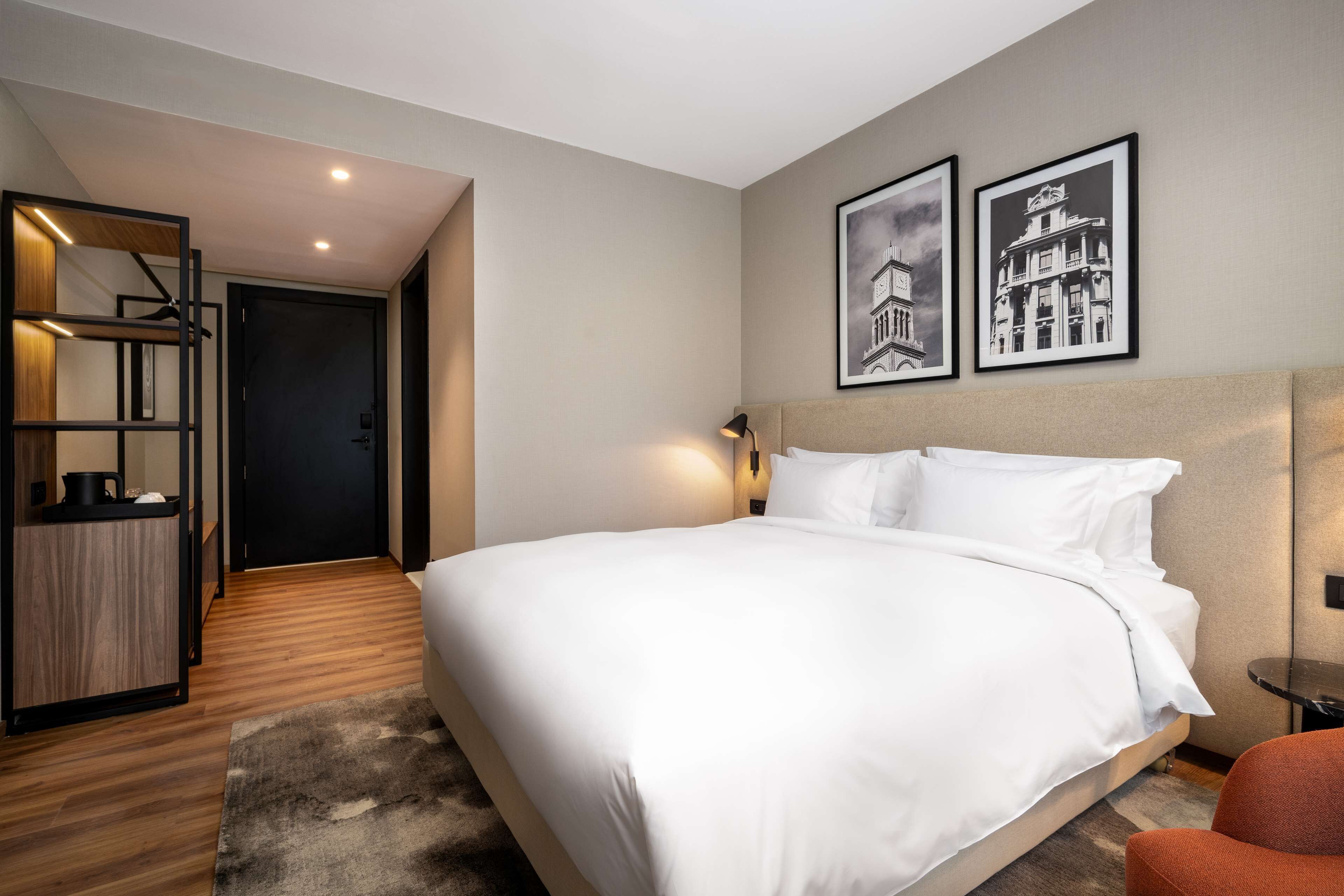 A modern hotel room with a large bed, a desk with a chair, and a wardrobe. There are two framed pictures on the wall.