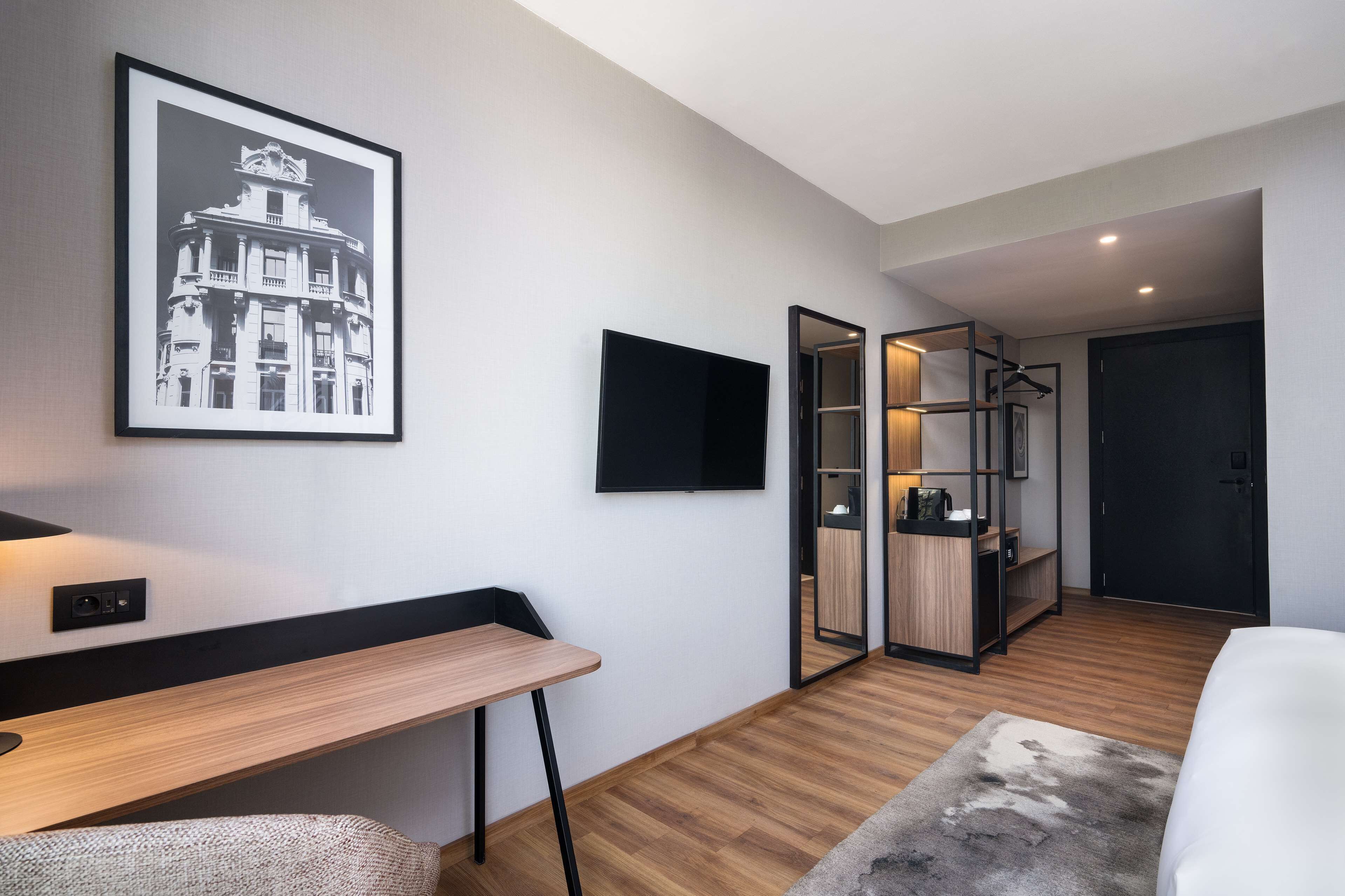 A modern hotel room with a wooden desk, a bed, a framed picture on the wall, a TV, and a walk-in closet.