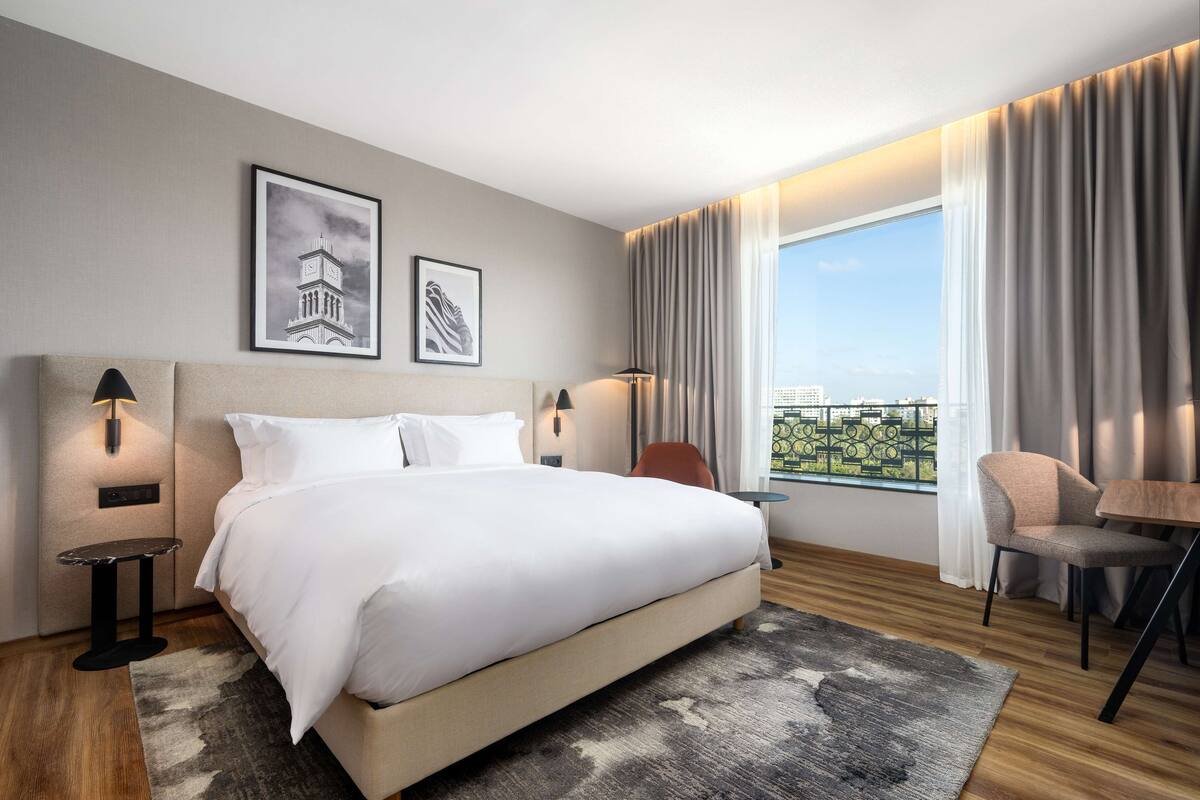 Une chambre d’hôtel moderne avec un grand lit, deux lampes fixées au mur, une petite table, une chaise et une vue sur la ville grâce à de grandes fenêtres.