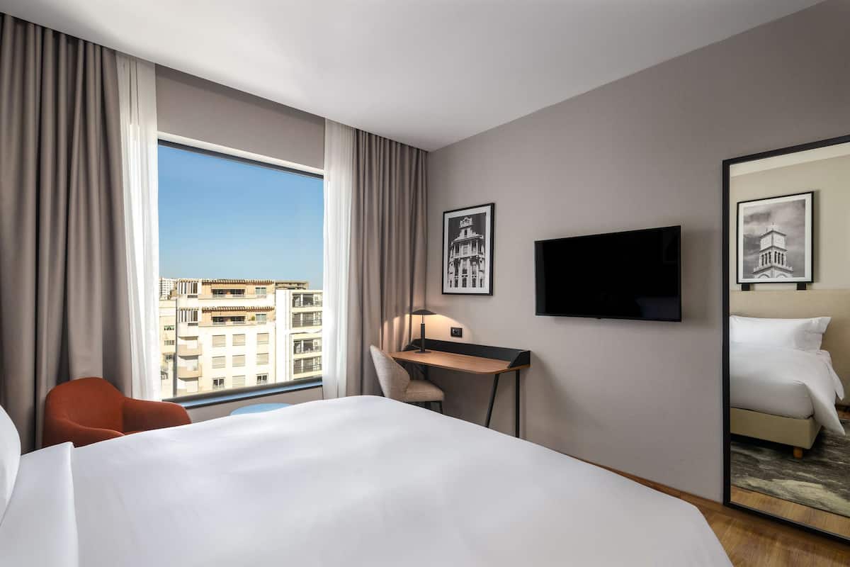 Une chambre d’hôtel avec un grand lit, un bureau, une chaise, une télévision et une fenêtre avec des rideaux.