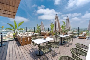 An outdoor patio with wicker furniture, white tables, and beige cushions, overlooking a cityscape.