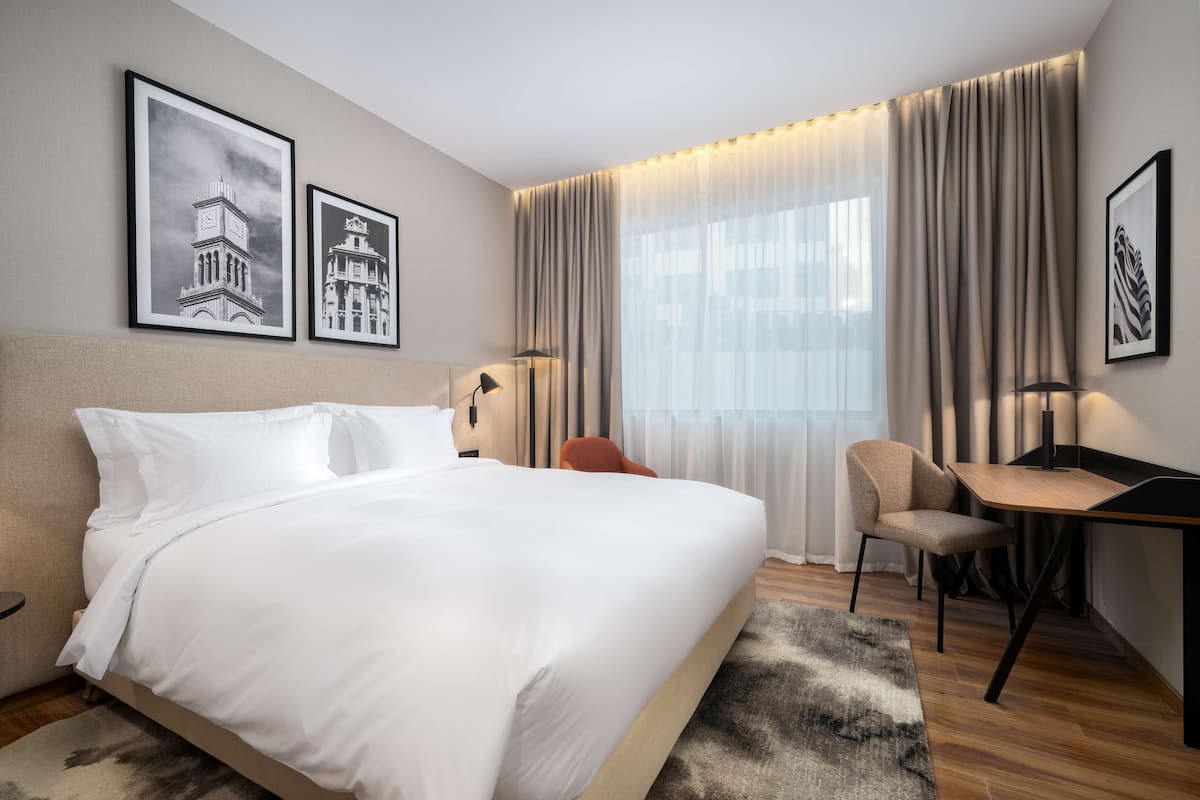 Une chambre d’hôtel moderne avec un grand lit, un bureau et deux chaises. Les murs sont ornés de tableaux encadrés et il y a une fenêtre avec des rideaux.