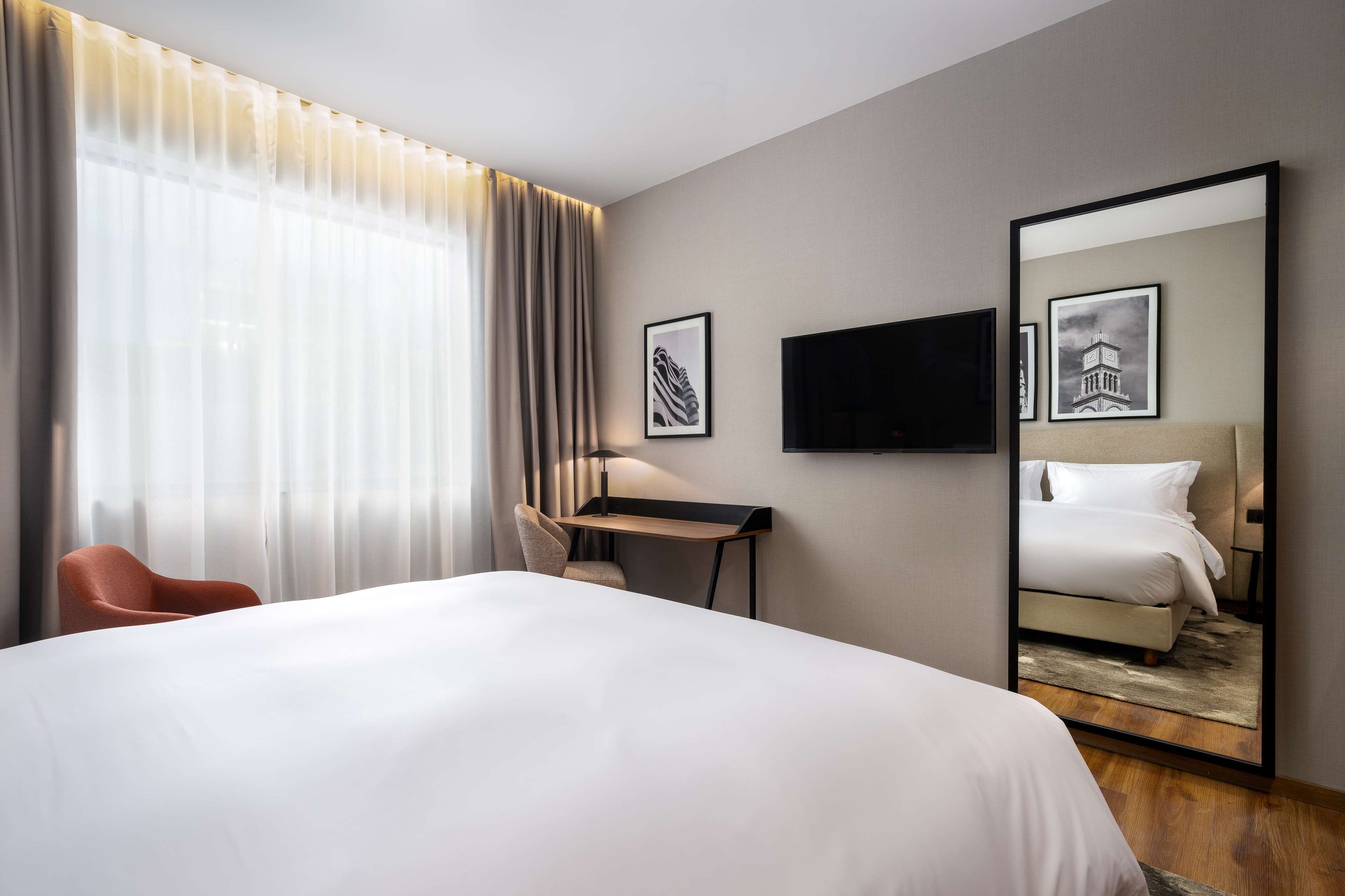 A hotel room with a large bed, a desk, a chair, a television, and a mirror reflecting the bed.
