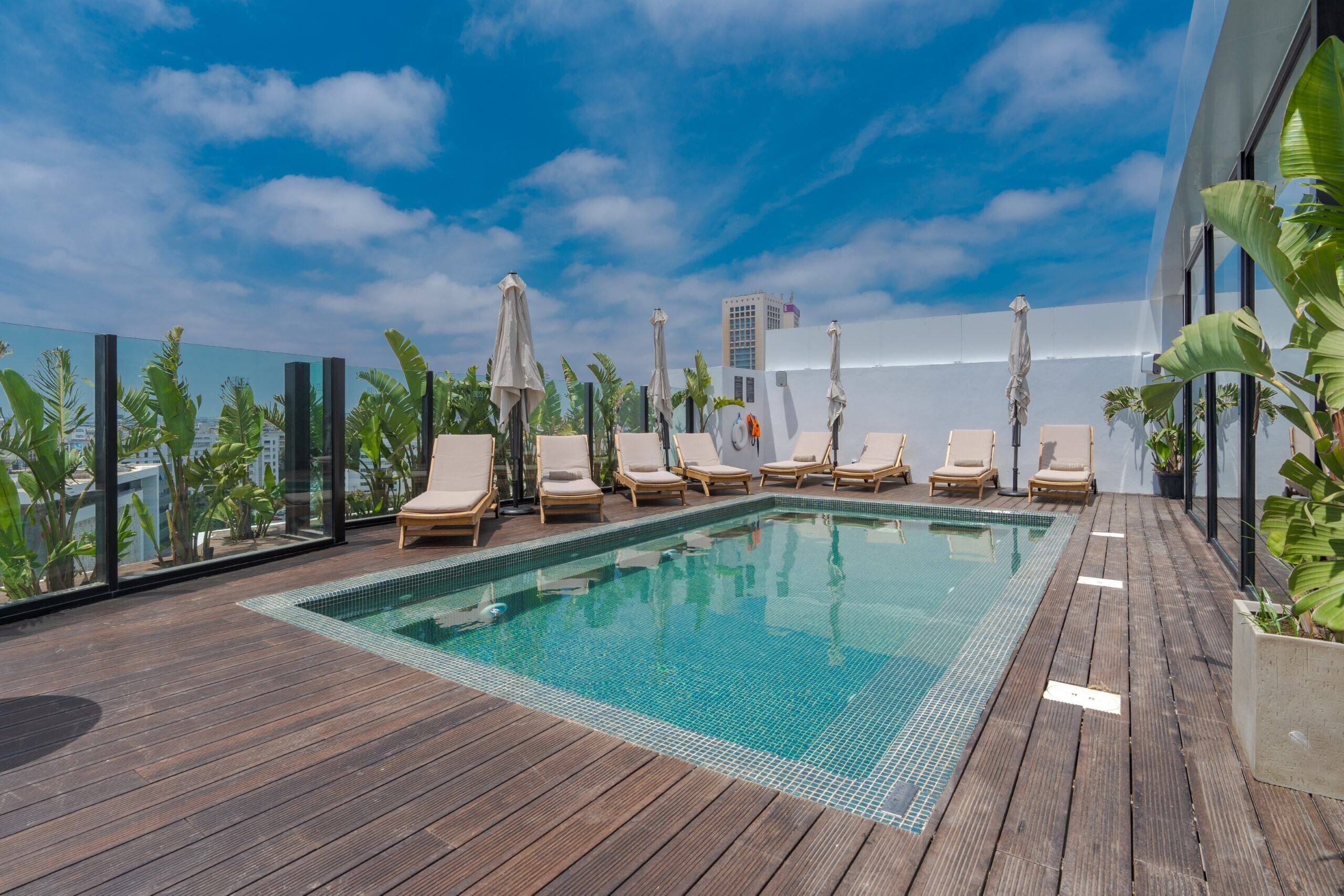 A rooftop pool area with lounge chairs and umbrellas, surrounded by glass walls and tropical plants.