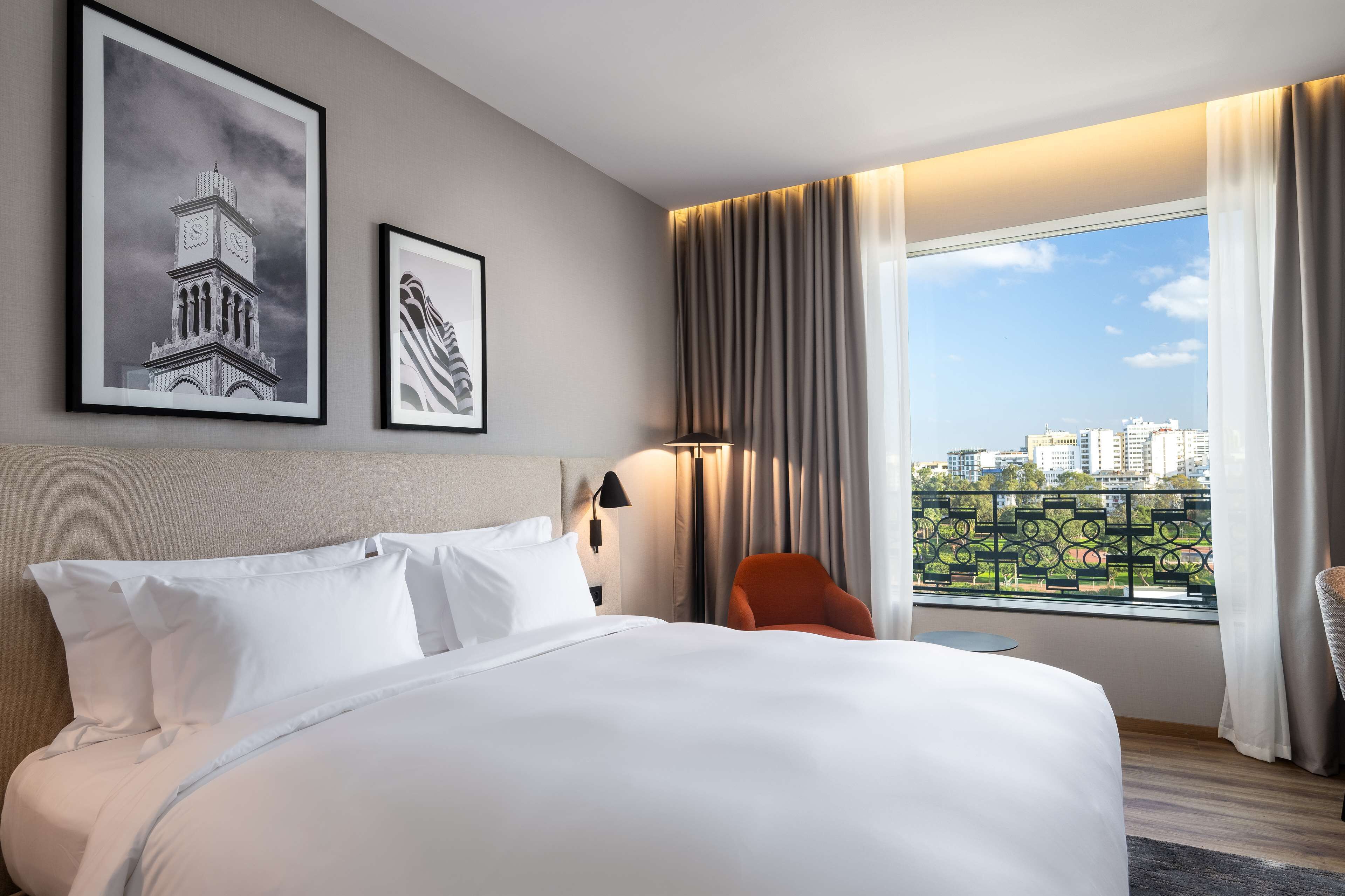 Habitación de hotel con una cama grande, una mesita de noche, una lámpara, una silla y una ventana con vistas a edificios.